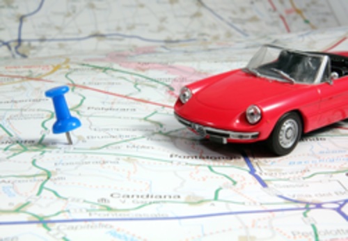 Ein rotes, kleines Auto fährt auf einer Landkarte, die mit einem Pin gekennzeichnet ist. Per GPS werden Ihre Fahrzeuge geortet