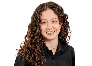 Porträt einer lächelnden Frau mit langen, lockigen braunen Haaren und schwarzer Bluse vor weißem Hintergrund.
