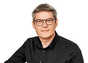 Porträt einer Person mit Brille und dunklem Hemd vor hellem Hintergrund